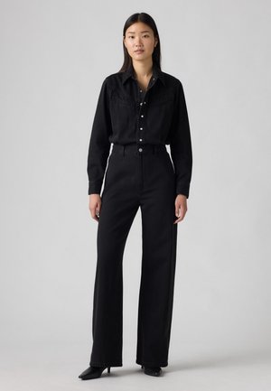 Chemise en denim noir avec empiècement style western, boutons-pression à l'avant, associée à un jean large taille haute noir et des talons aiguilles noirs.