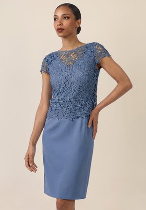 Frau trägt ein blaues Spitzenoberteil mit kurzen Ärmeln und Blumenmustern, kombiniert mit einem einfarbigen blauen knielangen Rock, steht vor einem beigen Hintergrund.