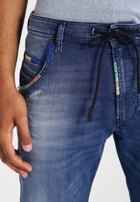 Primer plano de una persona que lleva unos vaqueros de mezclilla azul desgastados con acentos de hilo colorido y un cordón negro, con la mano descansando junto al muslo.