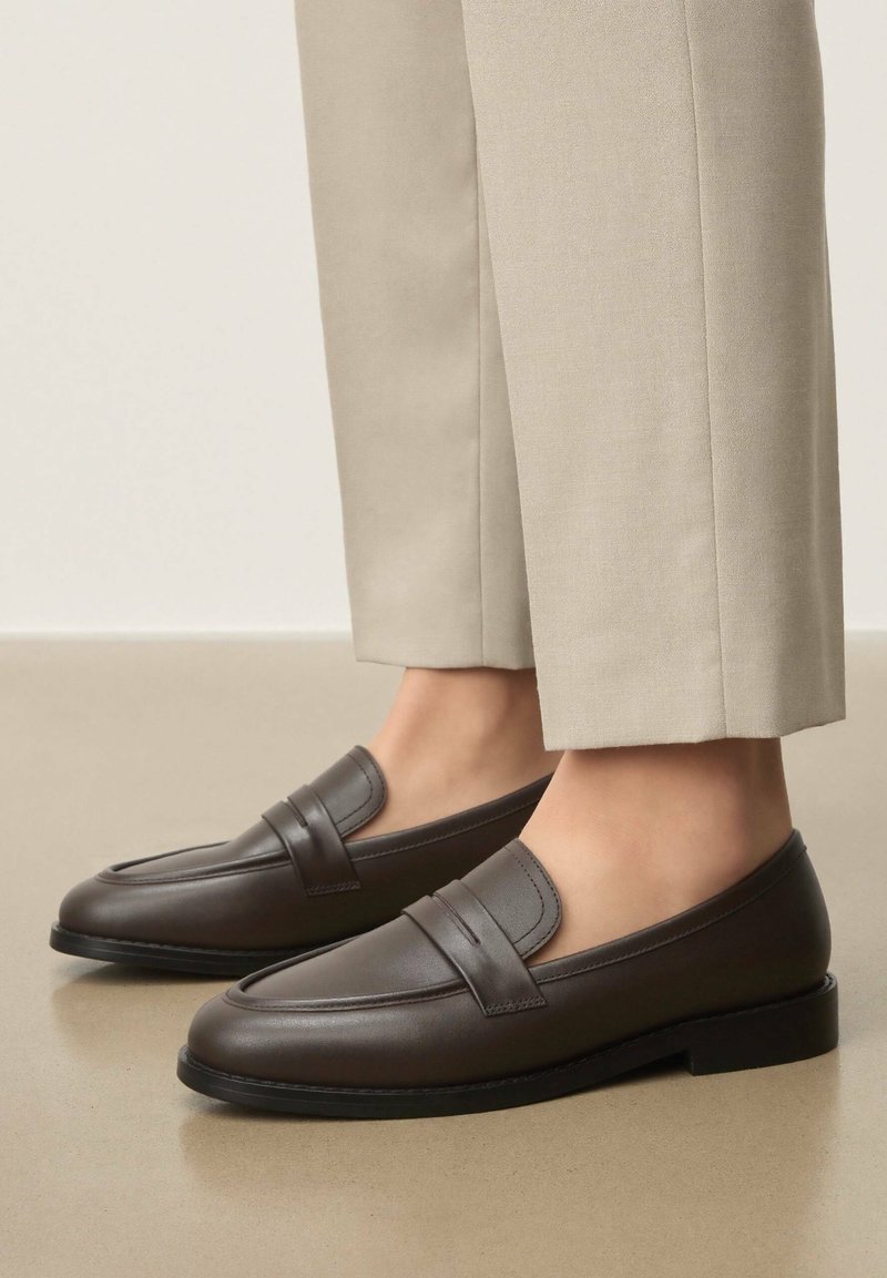 Mocassins en cuir marron à finition lisse, avec une couture légèrement relevée et un bout arrondi, associés à un pantalon beige clair.