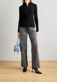 Schwarzes, geripptes Langarmtop mit Reißverschluss am Ausschnitt, kombiniert mit grauen Jeans mit weitem Bein und spitzen schwarzen High Heels. Hellblaue Handtasche mit glatter Oberfläche.