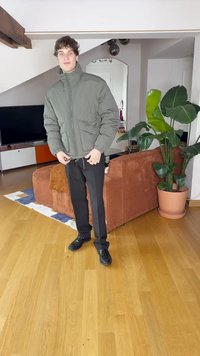 Padded olive green jacket with a high collar, black trousers with side stripes, and black shoes, set in a bright indoor environment.