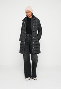 Czarna puchowa kurtka z zamkiem z przodu, przyszytym kapturem i zatrzaskami. W zestawie z czarnym swetrem i szerokimi szarymi dżinsami. Jasnoróżowa dziergana czapka.