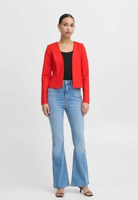 Červený zkrácený blazer s dlouhými rukávy a otevřeným předním designem. V kombinaci s vysokými modrými džiny a černými sandály na podpatku.