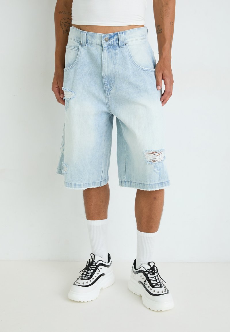 Shorts en denim bleu clair avec une coupe décontractée, présentant des détails déchirés et des ourlets effilochés. Associés à des chaussettes blanches et des baskets noir et blanc.