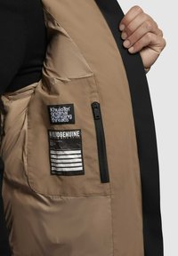 Hand die een beige jas vasthoudt, waarbij de binnenlabels en een te ritsen zak aan de binnenzijde links zichtbaar zijn.