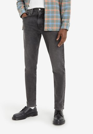 Person trägt schwarze, ausgewaschene Jeans, schwarze Schnürschuhe, weiße Socken, ein weißes Hemd und eine mehrfarbige karierten Jacke.