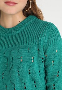 Stickad tröja i teal med ett strukturerat mönster som har vågiga designer och små utskärningar. Ribbad halsringning och mjukt, tjockt material.