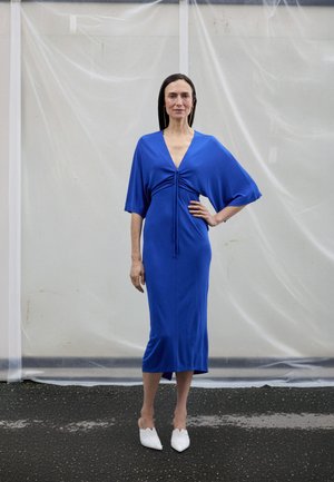 Blå knälång klänning med korta, vida ärmar och en rynkad mitt. Kombinerad med vita skor med spetsig tå och texturerad design.