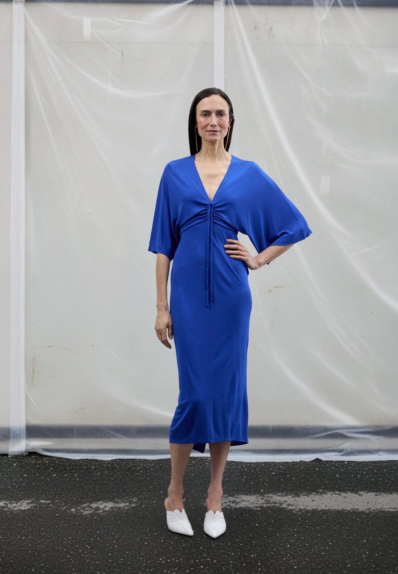 Modra obleka do kolen s kratkimi, širokimi rokavi in naborkom na sredini. Kombinirana z belimi čevlji z ostrim koncem in teksturirano zasnovo.