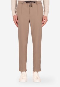 Pantaloni beige con vestibilità slim, dotati di vita con coulisse e pieghe frontali, abbinati a sneakers bianche. Tessuto liscio, dettagli minimi.