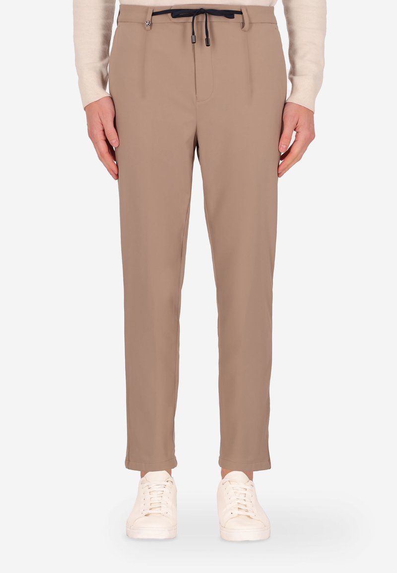 Pantaloni beige con vestibilità slim, dotati di vita con coulisse e pieghe frontali, abbinati a sneakers bianche. Tessuto liscio, dettagli minimi.
