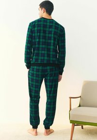 Grøn- og marineblå plaid flannel pyjamas sæt med rund halsudskæring, elastiske manchetter og talje. Blød tekstur. Barfodet, stående op ad en væg.
