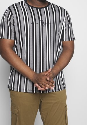 Uomo con le mani incrociate che indossa una maglietta a righe verticali bianche e nere e pantaloni cargo marroni, su sfondo bianco uniforme.