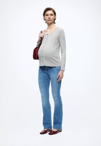 Svetlosivý rebrovaný kardigán s dlhými rukávmi na gombíky, skombinovaný s modrými rozšírenými rifľami a červenými balerínkami z lakovanej kože. Modelka drží červenú kabelku.