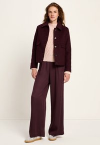 Woman stands wearing a dark purple buttoned jacket, light pink textured sweater, wide-legged purple pants, and white sneakers with gum soles.