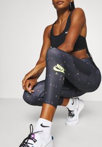 Atletka ve dřepu, oblečená v černé sportovní podprsence Nike, černých legínách Nike Air s barevným textem, bílých ponožkách Nike a bílých teniskách s černými tkaničkami.
