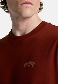 Camiseta de manga corta marrón con cuello redondo, que presenta un logo circular bordado en dorado en el pecho. Material de algodón suave.