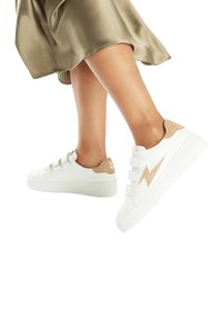 Baskets blanches avec une semelle en caoutchouc texturé et des accents en daim beige. Dotées d'un design en forme d'éclair et de bretelles élastiques pour un enfilage facile.