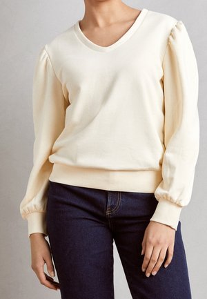 Femme portant un pull crème à col en V avec des manches longues bouffantes et un jean bleu foncé sur un fond uni.