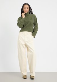 Pulover verde cu guler înalt, tricotat, cu modele din cabluri, asortat cu pantaloni largi de culoare crem și botine cu toc de culoare verde măslin.