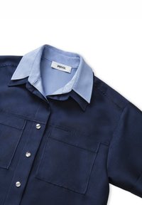 Navy blue shirt with a light blue collar, featuring two front pockets and silver snap buttons. Made of smooth fabric with a textured weave.