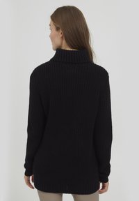 Pull en maille noire avec un col roulé côtelé haut, coupe ample et manches longues avec des poignets côtelés. Surface texturée visible depuis l'arrière.