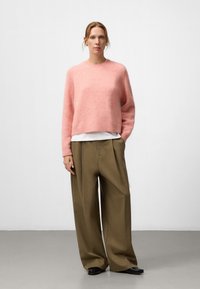Różowy, puszysty sweter z okrągłym dekoltem i ribbami u mankietów, w połączeniu z luźnymi, oliwkowymi spodniami z plisami oraz czarnymi butami.