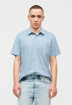 Νεαρός άνδρας με κοντά μαλλιά, φορώντας μπλε-λευκό καρό κοντομάνικο πουκάμισο, μερικώς μπουσουριάσμένο σε ανοιχτόχρωμο τζιν, στέκεται μπροστά σε απλό φόντο.