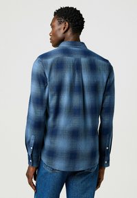 Camisa de cuadros de manga larga en tonos de azul, con cuello abotonado, textura sutil y puños de un solo botón. La vista trasera muestra un diseño de yugo.