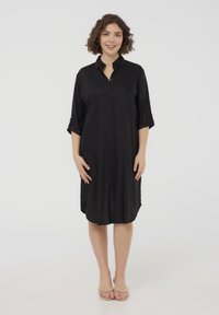 Abito camicia nera con bottoni, realizzato in un tessuto leggero, con maniche arrotolabili e orlo asimmetrico. Texture liscia e vestibilità rilassata.