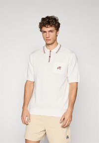 Hvit polo-skjorte med korte ermer, med krage i burgunderstriper, en brystlomme og et brodert logo. Glatt tekstur.