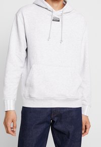Person som bär en ljusgrå Adidas-hoodie med en framficka och dragsnören, kombinerad med mörkblå jeans mot en enkel bakgrund.