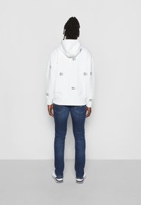 Witte hoodie met een herhaald patroon van zwarte tekst, verstelbare capuchon, geribbelde manchetten, gecombineerd met blauwe jeans en witte sneakers.