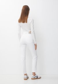 Žena se rovnými hnědými vlasy stojí čelem k bílé zdi, oblečena v bílém tričku s dlouhým rukávem, bílých džínách a bílých teniskách s černými akcenty.