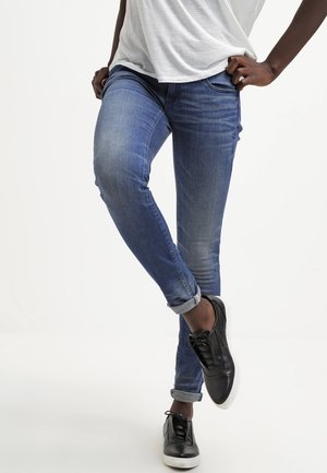 Personne portant un jean bleu retroussé, des baskets noires et une chemise blanche, debout avec une jambe croisée sur l'autre.