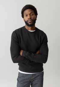 Felpa nera con scollo tondo, caratterizzata da maniche lunghe e una finitura texturizzata. Indossata sopra una camicia bianca e abbinata a jeans in denim scuri.