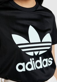 Mustattu lyhythihaisessa paidassa on sileä kangas, jossa on suuri valkoinen Adidas trefoil -logo ja brändin nimi vahvasti esillä.