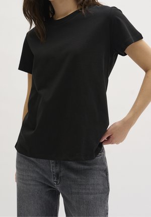 Person wearing a plain black short-sleeve t-shirt and gray jeans, hand resting on hip against a plain white background.