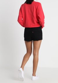 Sudadera roja con mangas raglán, pantalones cortos de denim negros con bordes deshilachados y zapatillas blancas, vista por la parte de atrás.