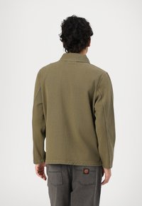Olive green cotton jacket with a structured design and long sleeves, featuring minimal stitching details and a branded patch near the hem.