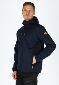 Mørkeblå softshell-jakke med hætte, der har to lynlåslommer på brystet og sidelommer, blevet fremhævet med brune detaljer og et emblem.