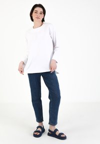Hvid langærmet top, afslappet pasform, parret med blå denim jeans og mørkeblå sandaler med justerbare stropper. Enkel og minimalistisk design.