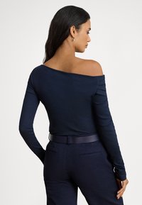 Hosszú sötét hajú nő, aki testhezálló sötétkék váll nélküli hosszú ujjú felsőt és hozzáillő öves nadrágot visel, háttal áll egy egyszínű háttér előtt.