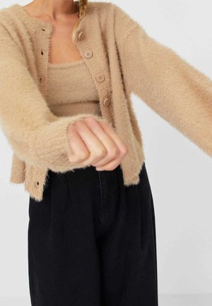 Femme portant un cardigan beige duveteux et un débardeur avec un pantalon noir, les bras tendus vers l'avant sur un fond clair uni.