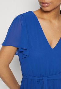 Femme portant une robe bleu royal à col en V avec des manches volantes superposées et une ceinture nouée à la taille, montrée de l'épaule à la poitrine.
