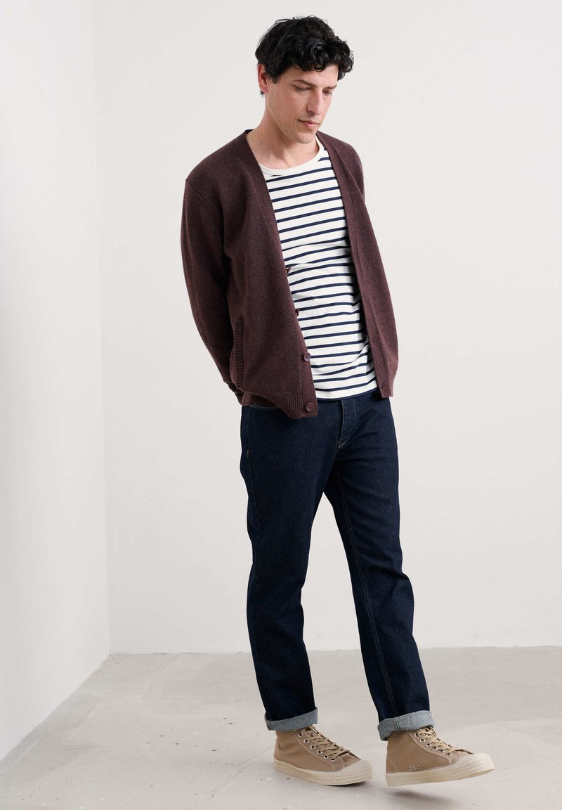 Maroon adītais džemperis virs balta un tumši zilā svītrainā T-kreklā, apvienots ar tumšiem džinsu biksēm un bēšiem augstiem sporta apaviem. Gluda tekstūra.