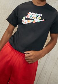 Osoba v černém tričku Nike s barevnými samolepkami s ovocným motivem na logu a jasně červených kalhotách na světle dřevěném pozadí.