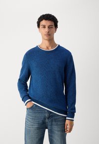 Blå strikkegenser med rund hals, med hvite ribbede detaljer på mansjettene og kanten. Brukt sammen med lyseblå denimjeans.