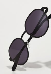 Ochelari de soare negri cu un cadru octagonal și lentile de culoare violet închis. Au un braț subțire, texturat, de culoare neagră și tampoane pentru nas transparente.